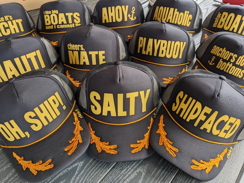May include: A collection of ten black trucker hats with gold embroidered text and designs. The hats feature various nautical-themed phrases such as "Ahoy", "Salty", "Shipfaced", "Oh, Ship!" and "Playbuoy".