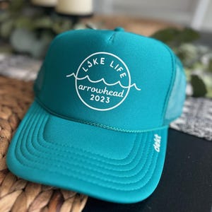 Puede incluir: Una gorra de camionero azul turquesa con un logotipo bordado en blanco que dice "Lake Life Arrowhead 2023".