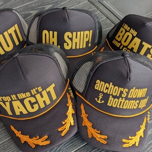 May include: Five black trucker hats with gold embroidery. The hats feature nautical themed phrases like "Nauti", "Oh Ship!" and "Anchors Down Bottoms Up!"