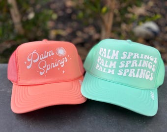 May include: Two trucker hats with the text "Palm Springs" in white on a pink and a green hat.