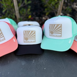 Puede incluir: Una colección de gorras de camionero en varios colores, incluyendo coral, verde menta y negro. Cada gorra presenta un panel frontal blanco con un diseño de rayos de sol dorado y el texto "CHASING THE SUN". Las gorras tienen una parte trasera de malla y una visera curva.