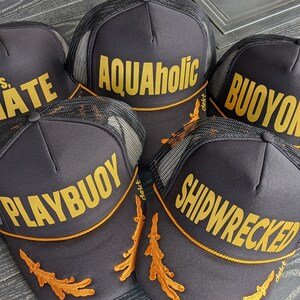 May include: Five black trucker hats with gold embroidered text. The hats feature different phrases including "Cheers, Mate", "Aquaholic", "Buoyonce", "Playbuoy", and "Shipwrecked".
