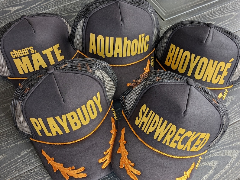 May include: Five black trucker hats with gold embroidered text. The hats feature different phrases including "Cheers, Mate", "Aquaholic", "Buoyonce", "Playbuoy", and "Shipwrecked".