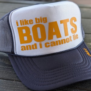 May include: A trucker hat with a white front panel and a gray mesh back. The front features the text "i like big BOATS and i cannot lie" in orange. The brim and the bottom of the hat are dark gray.
