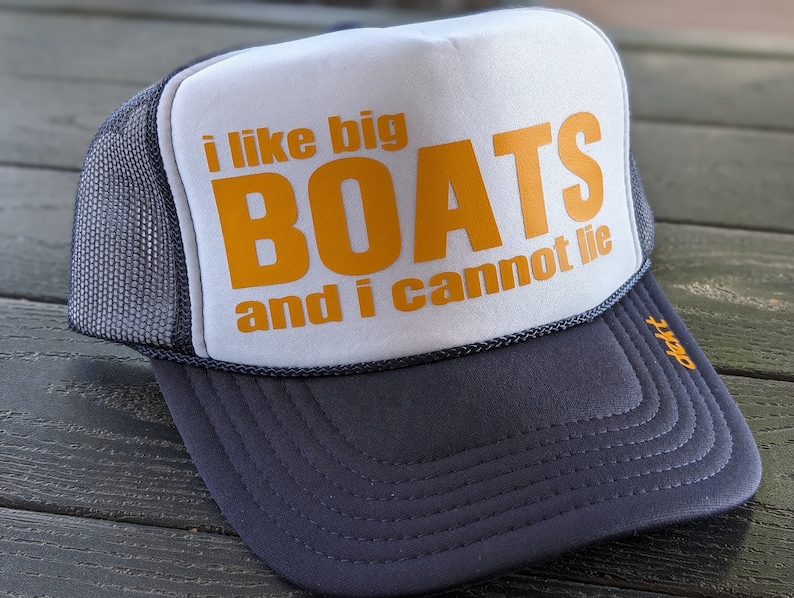 May include: A trucker hat with a white front panel and a gray mesh back. The front features the text "i like big BOATS and i cannot lie" in orange. The brim and the bottom of the hat are dark gray.