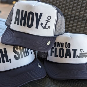 May include: Three navy blue and white trucker hats with nautical-themed phrases. One hat says "AHOY" with an anchor graphic, another says "OH, SHIP!", and the third says "down to FLOAT." Each hat has a mesh back.