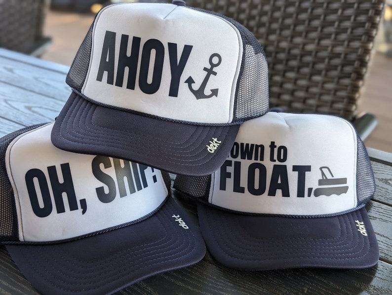 May include: Three navy blue and white trucker hats with nautical-themed phrases. One hat says "AHOY" with an anchor graphic, another says "OH, SHIP!", and the third says "down to FLOAT." Each hat has a mesh back.