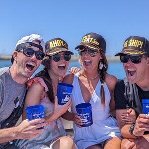 May include: Four friends on a boat wearing black and gold baseball caps with nautical themed text. The caps say "Ahoy", "Ready to Boat", and "Oh Ship!" The friends are holding blue plastic cups with the text "Ain't no party like a boat party".
