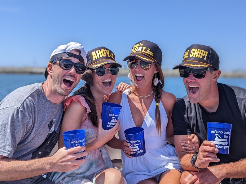 May include: Four friends on a boat wearing black and gold baseball caps with nautical themed text. The caps say "Ahoy", "Ready to Boat", and "Oh Ship!" The friends are holding blue plastic cups with the text "Ain't no party like a boat party".