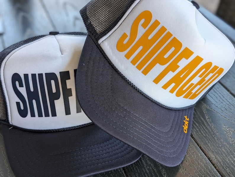 May include: Two trucker hats with white front panels and black mesh backs. One hat has the word "SHIPFACED" in black block letters, and the other has the same word in orange. Both hats have a dark gray brim.