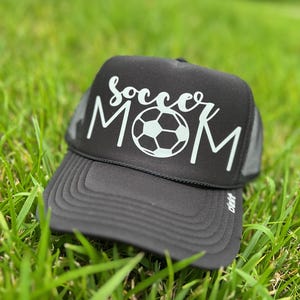 Puede incluir: Gorra de camionero negra con un balón de fútbol blanco y el texto "Soccer Mom" en blanco.