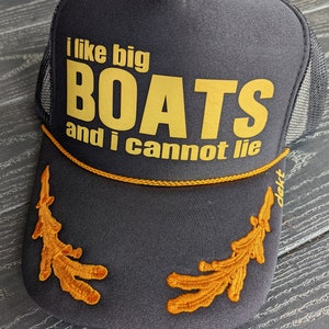 May include: A black trucker hat with a gold embroidered design. The hat has a mesh back and says "I like big boats and I cannot lie".