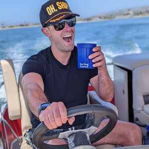 May include: A person wearing a black baseball cap with the text "OH, SHIP" in yellow letters, sunglasses, and a black t-shirt, is holding a blue cup with the text "Sea Dog Seamus" and driving a boat.