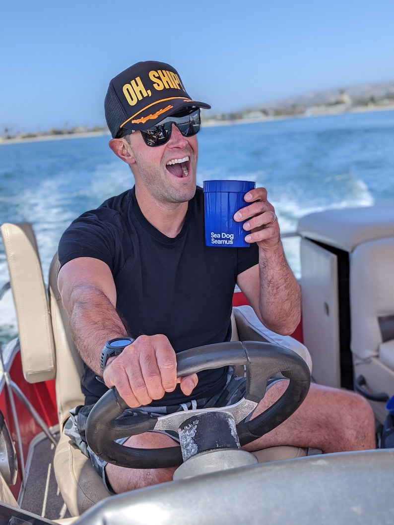 May include: A person wearing a black baseball cap with the text "OH, SHIP" in yellow letters, sunglasses, and a black t-shirt, is holding a blue cup with the text "Sea Dog Seamus" and driving a boat.