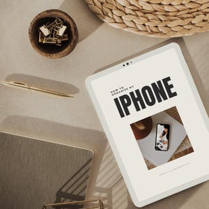 Puede incluir: Una tableta blanca con un fondo gris claro y el texto "HOW TO ORGANIZE MY IPHONE" en negro. La tableta está sobre una superficie marrón claro con una cesta de mimbre y un pequeño cuenco de clips de papel dorados en el fondo.