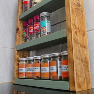 May include: A wooden spice rack with three shelves painted green. The shelves are filled with glass jars of spices, including ground ginger, ground cinnamon, ground nutmeg, ground coriander, and red chili powder. The jars have labels with the names of the spices and the brand "Sainsbury's".