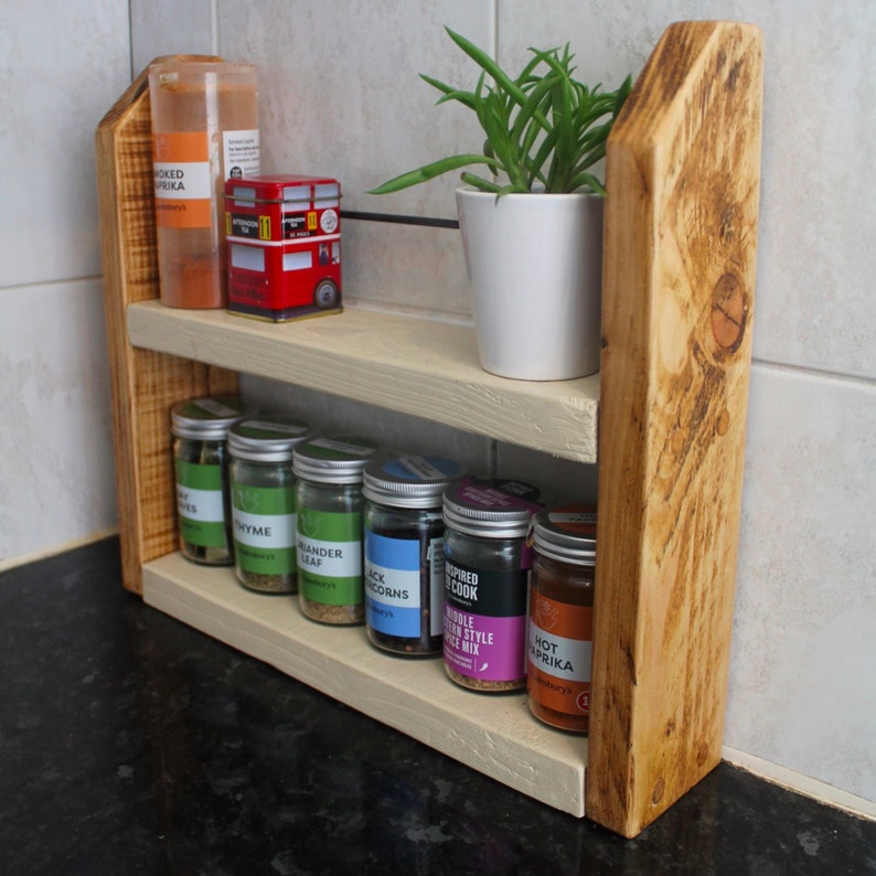 May include: A two-tiered wooden spice rack with a light brown finish. The top shelf has a red tin with a London bus design and a glass jar with a red label. The bottom shelf has seven glass jars with various labels, including "Thyme", "Black Peppercorns", and "Hot Paprika".