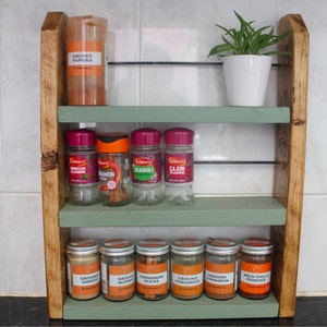 May include: A wooden spice rack with three shelves painted in a light green color. The top shelf has a jar of smoked paprika. The middle shelf has four glass jars of spices labeled "Moroccan Seasoning", "Cinnamon Sticks", "Seasoned", and "Cajun Seasoning". The bottom shelf has seven glass jars of spices labeled "Ground Ginger", "Ground Nutmeg", "Cinnamon Sticks", "Ground Coriander", "Ground Cinnamon", and "Mild Chilli Powder".