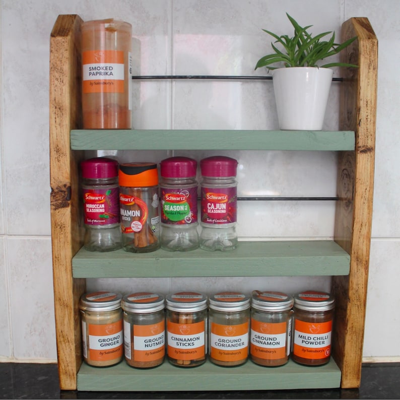 May include: A wooden spice rack with three shelves painted in a light green color. The top shelf has a jar of smoked paprika. The middle shelf has four glass jars of spices labeled "Moroccan Seasoning", "Cinnamon Sticks", "Seasoned", and "Cajun Seasoning". The bottom shelf has seven glass jars of spices labeled "Ground Ginger", "Ground Nutmeg", "Cinnamon Sticks", "Ground Coriander", "Ground Cinnamon", and "Mild Chilli Powder".