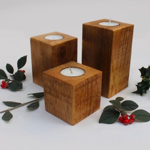 May include: Three wooden candle holders of varying sizes, each with a white tea light candle inside. The holders are made of rustic wood with a natural brown finish. The candles are arranged on a white surface with green leaves and red berries scattered around them.