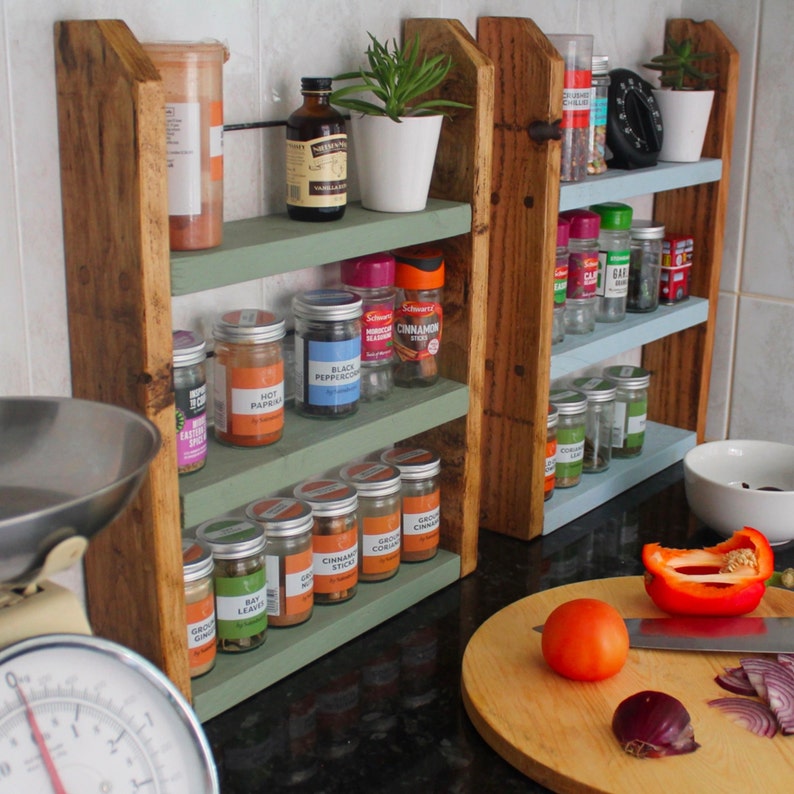 May include: A wooden spice rack with three shelves, painted in green and blue. The shelves are filled with various spices and seasonings in glass jars. The rack is mounted on a wall and has a rustic, farmhouse style.