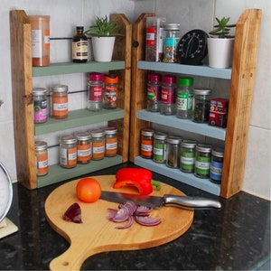 May include: A wooden spice rack with three shelves, painted in light green and blue. The rack is filled with various spices and herbs in glass jars. The rack is sitting on a black countertop next to a wooden cutting board with a red pepper, a red onion, and a knife.