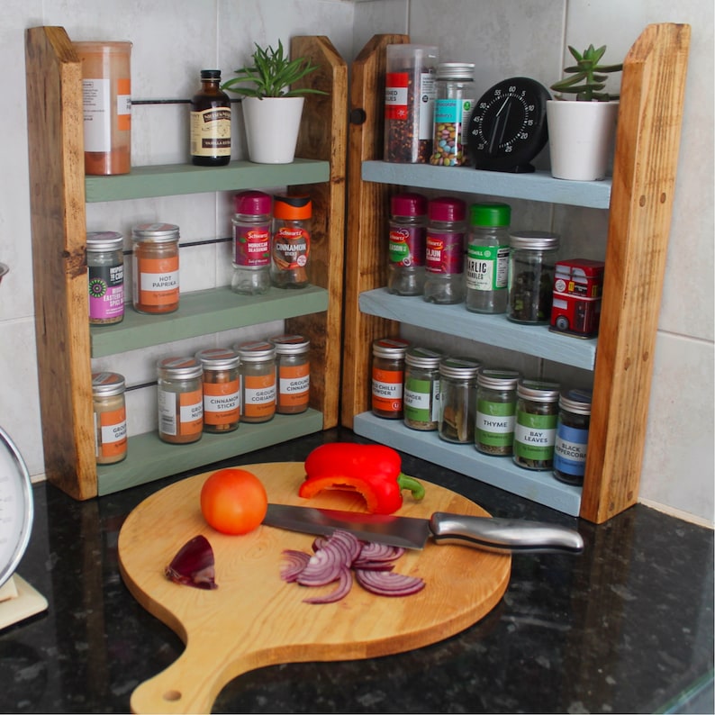 May include: A wooden spice rack with three shelves, painted in light green and blue. The rack is filled with various spices and herbs in glass jars. The rack is sitting on a black countertop next to a wooden cutting board with a red pepper, a red onion, and a knife.