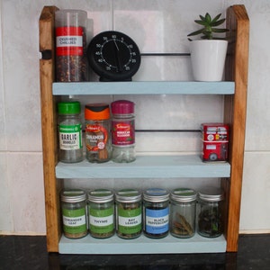 May include: A wooden spice rack with three shelves, painted light blue. The top shelf has a small white pot with a green succulent plant. The middle shelf has three glass spice jars with labels: "Garlic Granules", "Cinnamon Sticks", and "Moroccan Seasoning". The bottom shelf has six glass spice jars with labels: "Coriander Leaf", "Thyme", "Bay Leaves", "Black Peppercorns", "Salt", and "Pepper".