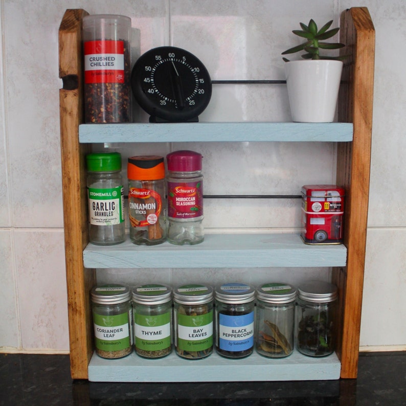 May include: A wooden spice rack with three shelves, painted light blue. The top shelf has a small white pot with a green succulent plant. The middle shelf has three glass spice jars with labels: "Garlic Granules", "Cinnamon Sticks", and "Moroccan Seasoning". The bottom shelf has six glass spice jars with labels: "Coriander Leaf", "Thyme", "Bay Leaves", "Black Peppercorns", "Salt", and "Pepper".