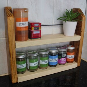 May include: A wooden spice rack with two shelves. The top shelf has a red tin that says "Afternoon Tea" and a glass jar with a label that says "Smoked Paprika". The bottom shelf has seven glass jars with labels that say "Bay Leaves", "Thyme", "Coriander Leaf", "Black Peppercorns", "Inspired to Cook Middle Eastern Spice Mix", and "Hot Paprika".