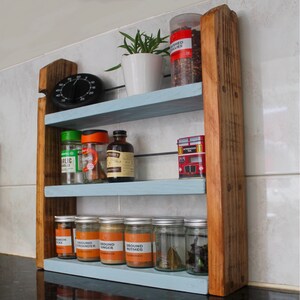 May include: A rustic wooden spice rack with three shelves painted light blue. The rack is filled with various spices in glass jars and tins. The top shelf has a small potted plant and a tin of dried chili peppers. The rack is mounted on a white tiled wall.
