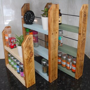 May include: Three wooden spice racks with three shelves each. The racks are painted in different colors: white, light blue, and sage green. The shelves are filled with various spices and herbs in glass jars. A small plant is on the top shelf of the white rack.
