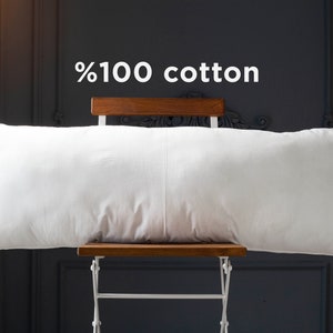 May include: A white, rectangular pillow made of 100% cotton, resting on a wooden chair against a dark gray wall.