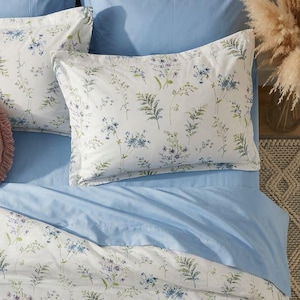 May include: A bed set featuring a light blue fitted sheet and pillowcases, with a white duvet cover and pillowcases adorned with a floral pattern in blue, green, and purple. A round, pink decorative pillow adds a contrasting texture.
