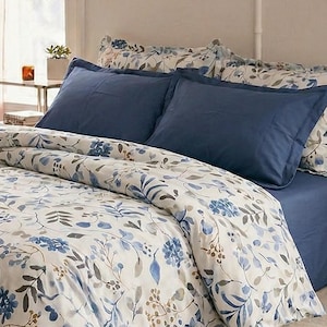 May include: A bed dressed with a floral duvet cover and blue sheets and pillowcases. The duvet cover has a white base with blue and grey floral and leaf patterns. The room features a green rug and a pink curtain.