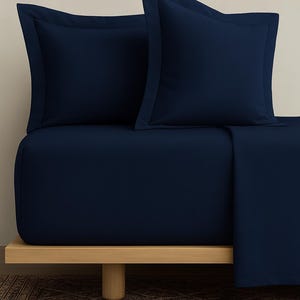 May include: A navy blue bed set, including two pillowcases and a fitted sheet, is displayed on a light wood bed frame. The pillowcases have a contrasting border. The bedding is neatly arranged, showcasing a minimalist aesthetic.