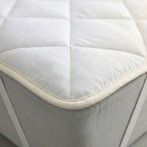 Puede incluir: Un protector de colch&oacute;n blanco con un patr&oacute;n acolchado en forma de diamante y un borde de tela color crema. El protector se asegura a un colch&oacute;n gris con correas el&aacute;sticas blancas en las esquinas que envuelven los bordes para fijar la funda.