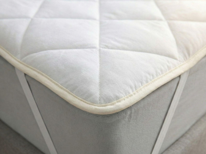 Puede incluir: Un protector de colch&oacute;n blanco con un patr&oacute;n acolchado en forma de diamante y un borde de tela color crema. El protector se asegura a un colch&oacute;n gris con correas el&aacute;sticas blancas en las esquinas que envuelven los bordes para fijar la funda.
