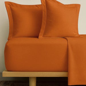 May include: An orange bed set with two pillowcases and a fitted sheet. The pillowcases have a contrasting border. The bed set is displayed on a wooden bed frame. The color is a warm, inviting shade.