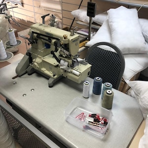 May include: A white industrial sewing machine with spools of thread in various colors, including white, blue, and green. The machine is on a table with a clear plastic container of sewing supplies. There is a business card on the table that says "KAYA HOME FURNISHINGS".