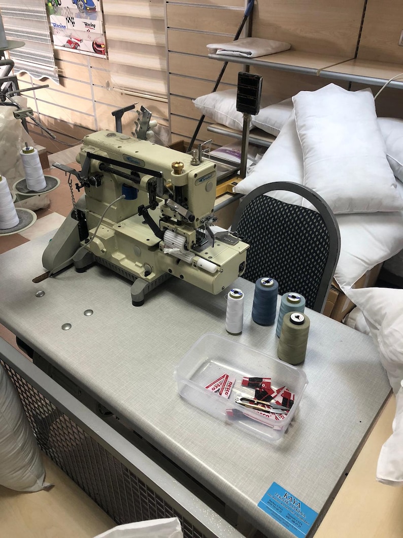 May include: A white industrial sewing machine with spools of thread in various colors, including white, blue, and green. The machine is on a table with a clear plastic container of sewing supplies. There is a business card on the table that says "KAYA HOME FURNISHINGS".