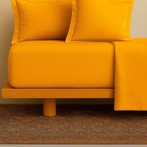 Peut inclure: Un lit avec un drap-housse jaune moutarde, deux taies d'oreiller assorties et une couverture pliée. Le lit repose sur un cadre en bois avec un seul pied cylindrique. Le fond est beige neutre.