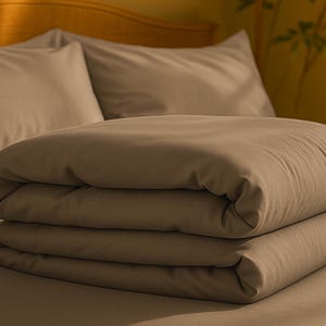 May include: A neatly folded set of taupe-colored bed linens, including a duvet and pillowcases, rests on a bed. The neutral color palette and soft textures suggest a focus on comfort and relaxation, ideal for bedroom decor.