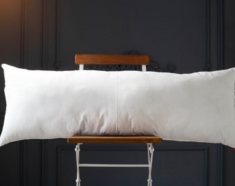 May include: A white rectangular pillow resting on a wooden chair with white metal legs. The pillow is against a dark grey background.
