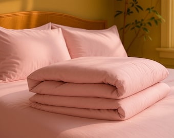 May include: A bed is made with light pink bedding. The set includes a fitted sheet, two pillowcases, and a folded comforter. The comforter is stacked on top of the bed. The headboard is made of wood.