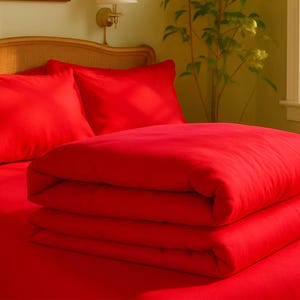 May include: A bed with red bedding, including two pillows and a folded blanket. The bed has a woven headboard and is in a room with a window and a wall-mounted lamp.