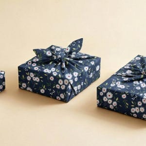 Può includere: Tre regali quadrati avvolti in tessuto blu navy con un motivo floreale bianco. Il tessuto è annodato sulla parte superiore, creando un fiocco decorativo. I regali sono disposti su una superficie beige chiaro.