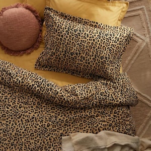 Könnte beinhalten: Ein Bettwäsche-Set mit einem Leopardenmuster-Bettbezug und Kissenbezügen. Die Bettwäsche liegt auf einem senfgelben Laken. Ein rundes, gefranstes, rosa Kissen und eine beige Decke sind ebenfalls zu sehen.