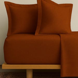 May include: A bed set featuring two square pillowcases and a fitted sheet in a warm, earthy orange-brown color. The pillowcases have a subtle border detail. The bedding is neatly arranged on a wooden bed frame, creating a cozy and inviting aesthetic.