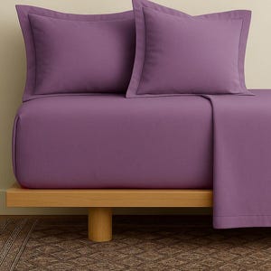 May include: A bed with a purple sheet set, including two pillowcases and a fitted sheet. The bedding is a solid color with a subtle border on the pillowcases. The bed frame is made of light-colored wood, and the rug below has a patterned design.